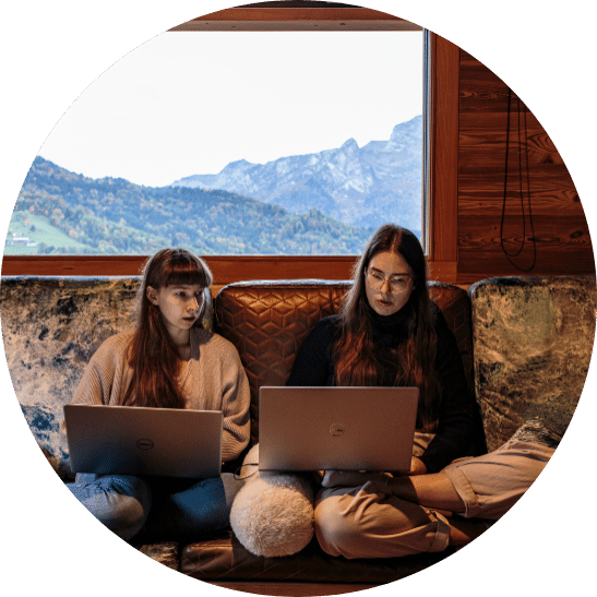 Zwei Frauen sitzen auf einem Sofa und schauen gemeinsam in ihre Laptops